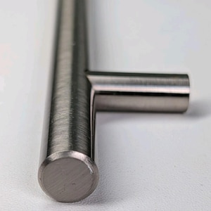 May include: A T-shaped cabinet handle with a brushed nickel finish. The hardware features a cylindrical bar design with a perpendicular mounting post extending from the center. The metallic surface displays a fine, linear texture throughout the piece.