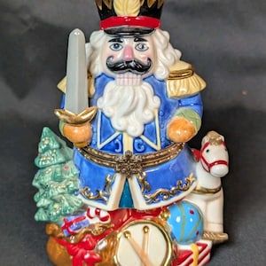 May include: A detailed ceramic Christmas nutcracker figurine. The figurine is dressed in a blue coat with gold accents, a black hat, and a white beard. It holds a sword and stands on a base with a Christmas tree, drum, and a toy horse.