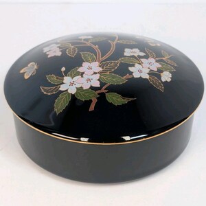 May include: A black, round decorative box with a glossy finish. The lid features a floral design with pink and white blossoms, green leaves, and a gold butterfly. The box has a gold trim around the base and lid.