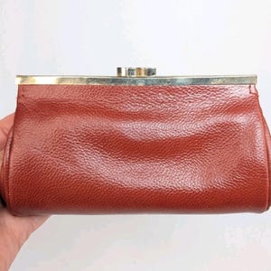 May include: A vintage, rust-colored leather clutch with a textured surface. The rectangular bag features a gold-toned metal clasp and frame. The clutch is held in a hand against a white background.