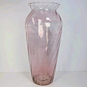 May include: A tall, translucent light pink glass vase featuring a subtle spiral or twisted rib pattern throughout its body. The vase has a wide, rounded base that tapers slightly toward the top, ending in a delicate, ruffled, and scalloped rim.