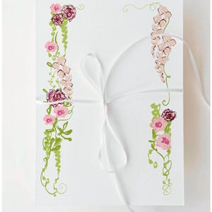Könnte beinhalten: Weißes Schreibwarenset mit floralen Illustrationen. Das Design zeigt rosa und burgunderfarbene Blumen mit grünen Ranken. Ein weißes Band ist um das Set gebunden und verleiht ihm eine zarte Note. Die Gesamtästhetik ist elegant und für verschiedene Anlässe geeignet.