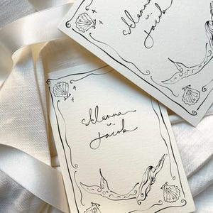 Pu&ograve; includere: Due biglietti color avorio con illustrazioni a inchiostro nero e calligrafia. I biglietti presentano un design a sirena, conchiglie e i nomi "Alanna & Jacob". I biglietti sono posizionati su un nastro di raso bianco.