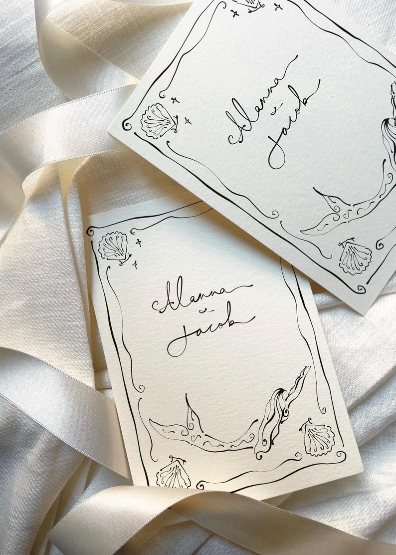 Pu&ograve; includere: Due biglietti color avorio con illustrazioni a inchiostro nero e calligrafia. I biglietti presentano un design a sirena, conchiglie e i nomi "Alanna & Jacob". I biglietti sono posizionati su un nastro di raso bianco.