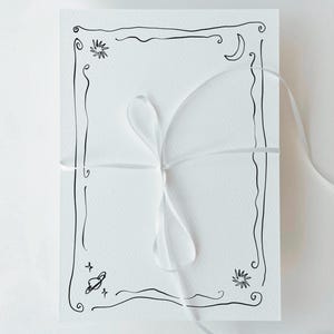 May include: A stack of white cards tied with a white ribbon. The cards have a black line drawing border with celestial elements like stars, a moon, and a planet. The overall aesthetic is minimalist and artistic.