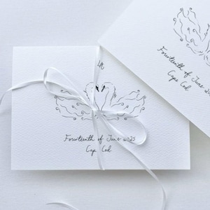 May include: White invitation cards with a swan illustration and the text "Fourteenth of June '25, Cape Cod". The cards are tied with a white ribbon, creating a delicate and elegant presentation. Ideal for weddings or special events.