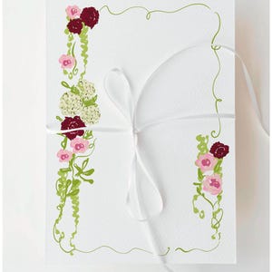 Könnte beinhalten: Weiße Karte mit floralen Illustrationen in Rosa, Burgunder und Creme, akzentuiert mit grünen Ranken. Ein weißes Band ist um die Karte gebunden. Das florale Design befindet sich auf der linken und rechten Seite der Karte, und eine grüne Rankenbordüre umgibt die Karte.
