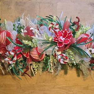 May include: A festive Christmas table runner featuring red poinsettias, striped ornaments, and white ribbon accents. The arrangement includes green foliage and glittery elements, creating a holiday centerpiece.
