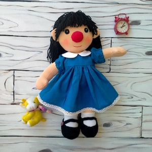 Pode incluir: Uma boneca de pano com cabelo de lã preta, nariz vermelho brilhante e bochechas rosadas, vestindo um vestido azul com gola branca e acabamento em renda, meias brancas e sapatos pretos. Uma pequena vaca de pelúcia amarela e um despertador analógico vermelho estão colocados por perto.