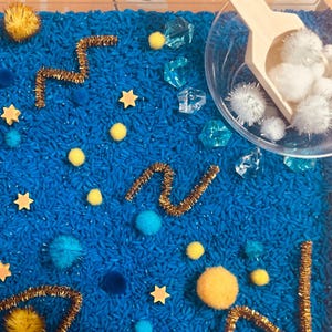 May include: A sensory bin filled with blue dyed rice, gold pipe cleaners, yellow pom-poms, star-shaped wooden cutouts, blue and white fuzzy balls, and clear plastic gems. A small wooden scoop and clear plastic bowl are visible.