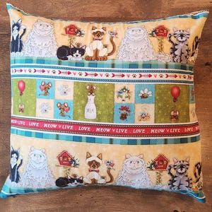 Pu&ograve; includere: Un cuscino decorativo con sfondo giallo e un motivo colorato di gatti, uccelli e case. Il cuscino ha un bordo a strisce blu, verde e rosso con il testo "MEOW &hearts; LIVE - LOVE - MEOW &hearts; LIVE - LOVE - MEOW &hearts; LIVE - LOVE" ripetuto in basso.