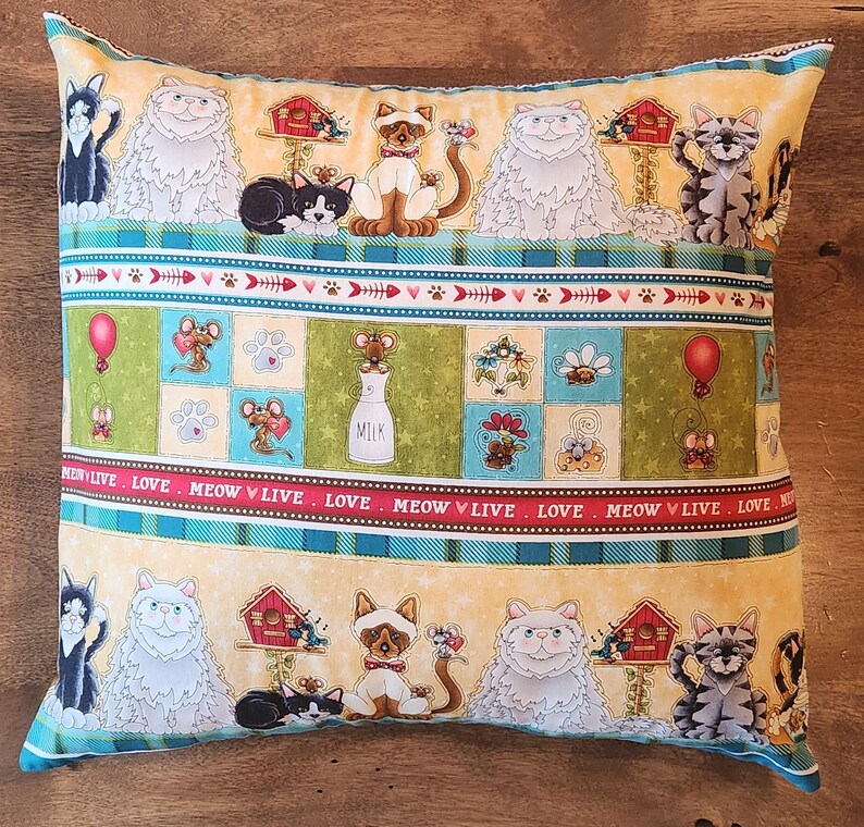 Pu&ograve; includere: Un cuscino decorativo con sfondo giallo e un motivo colorato di gatti, uccelli e case. Il cuscino ha un bordo a strisce blu, verde e rosso con il testo "MEOW &hearts; LIVE - LOVE - MEOW &hearts; LIVE - LOVE - MEOW &hearts; LIVE - LOVE" ripetuto in basso.