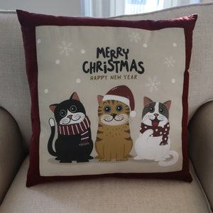 Pu&ograve; includere: Un cuscino rosso e beige a tema natalizio. Il cuscino presenta tre gatti dei cartoni animati che indossano cappelli e sciarpe invernali. Il testo sul cuscino recita "Merry Christmas Happy New Year".