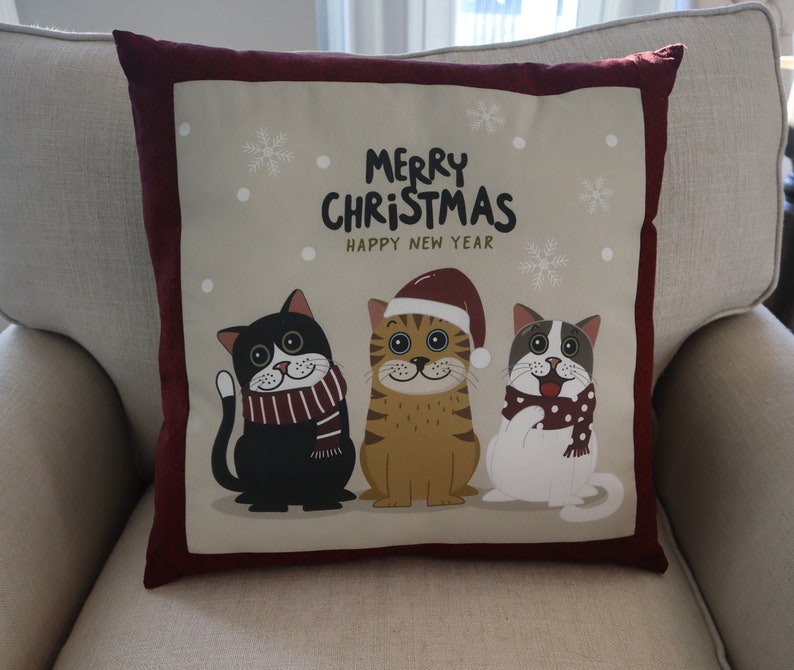 Pu&ograve; includere: Un cuscino rosso e beige a tema natalizio. Il cuscino presenta tre gatti dei cartoni animati che indossano cappelli e sciarpe invernali. Il testo sul cuscino recita "Merry Christmas Happy New Year".