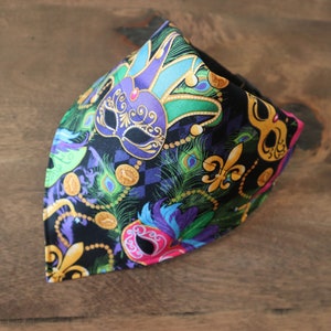 May include: A black bandana with a Mardi Gras theme. The bandana features purple, green, and gold masks, fleur-de-lis, and peacock feathers.