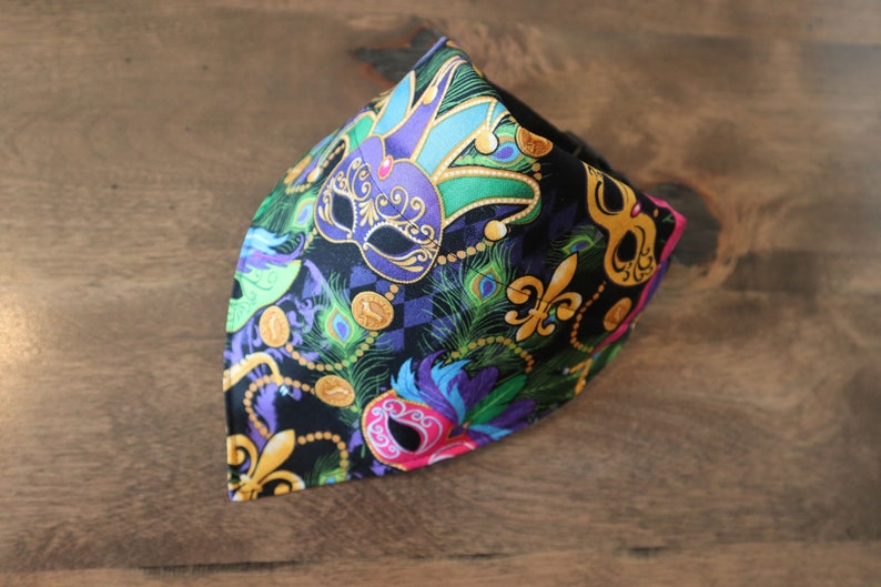May include: A black bandana with a Mardi Gras theme. The bandana features purple, green, and gold masks, fleur-de-lis, and peacock feathers.