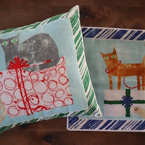 Pu&ograve; includere: Due cuscini decorativi con sfondo a righe blu e bianco. Un cuscino presenta un gatto grigio seduto su una scatola regalo rossa e bianca con cerchi bianchi. L'altro cuscino presenta un gatto arancione seduto su una scatola regalo verde e bianca con una stella blu.