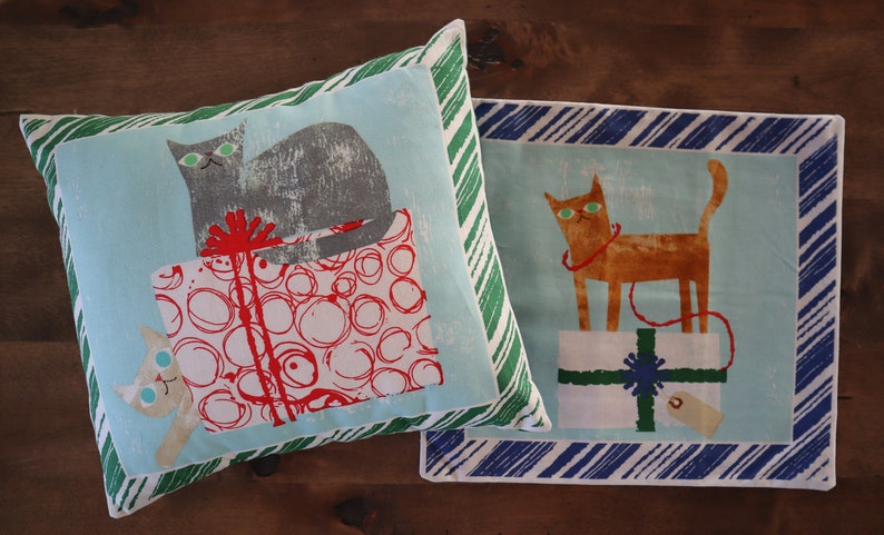 Pu&ograve; includere: Due cuscini decorativi con sfondo a righe blu e bianco. Un cuscino presenta un gatto grigio seduto su una scatola regalo rossa e bianca con cerchi bianchi. L'altro cuscino presenta un gatto arancione seduto su una scatola regalo verde e bianca con una stella blu.