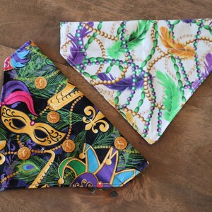 May include: Two fabric dog bandanas with Mardi Gras themed prints. One bandana is black with gold, green, and purple Mardi Gras beads, a jester's hat, and a mask. The other bandana is white with gold, green, and purple Mardi Gras beads and feathers.