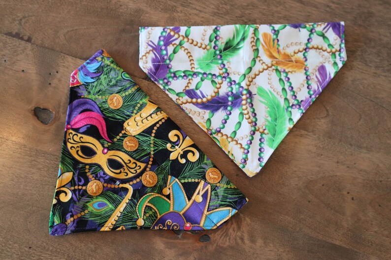 May include: Two fabric dog bandanas with Mardi Gras themed prints. One bandana is black with gold, green, and purple Mardi Gras beads, a jester's hat, and a mask. The other bandana is white with gold, green, and purple Mardi Gras beads and feathers.