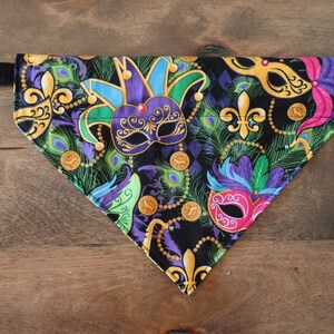 May include: A black fabric dog bandana with a Mardi Gras theme. The bandana features colorful masks, feathers, and gold beads on a black background. The bandana has a black adjustable strap.