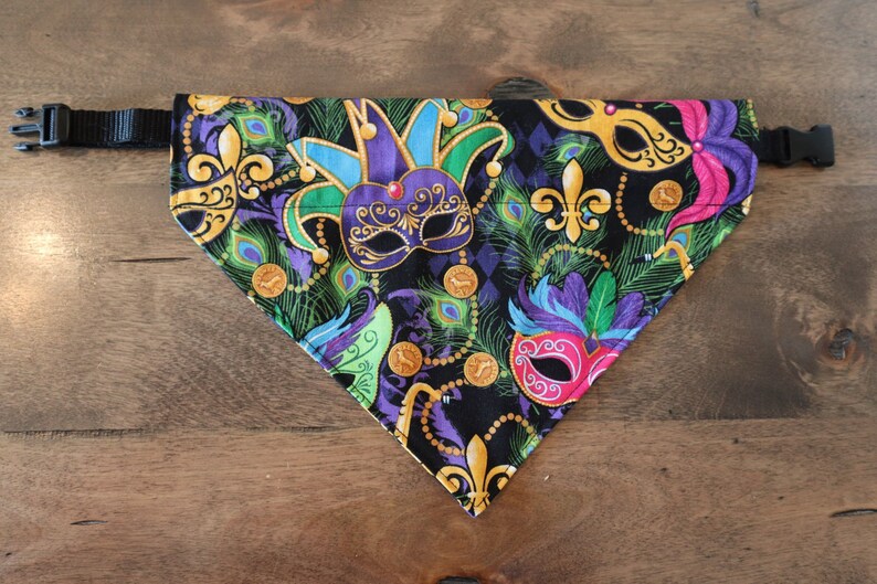 May include: A black fabric dog bandana with a Mardi Gras theme. The bandana features colorful masks, feathers, and gold beads on a black background. The bandana has a black adjustable strap.