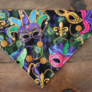 May include: A black fabric dog bandana with a Mardi Gras theme. The bandana features colorful masks, gold beads, and green, purple, and gold feathers.