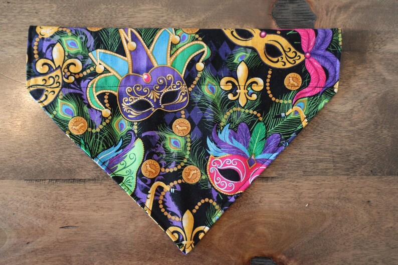 May include: A black fabric dog bandana with a Mardi Gras theme. The bandana features colorful masks, gold beads, and green, purple, and gold feathers.