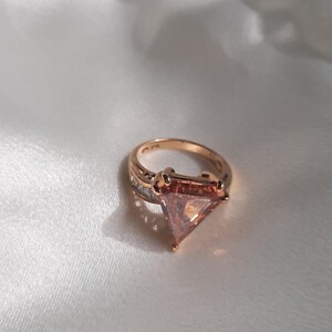 May include: A gold ring with a large, triangular, rose-colored gemstone surrounded by smaller clear gemstones.