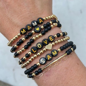 May include: A stack of black and gold beaded bracelets with the words "MAMA", "LOVE", and "SAM" spelled out in gold letters. The bracelets are on a person's wrist.