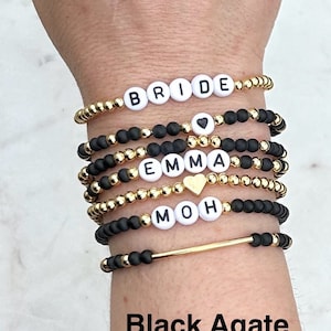 May include: A collection of beaded bracelets in gold and black. Some bracelets spell out "BRIDE", "EMMA", and "MOH" with white letter beads. Other bracelets feature heart-shaped beads. The text "Black Agate" is at the bottom.