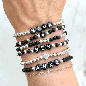 May include: A stack of black and silver bead bracelets with the names "MAMA", "BEN", and "ANNA" spelled out in white letters. Each bracelet has a small silver heart charm.