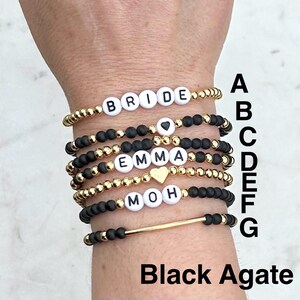 May include: A stack of seven beaded bracelets featuring gold-tone metallic beads, matte black beads, and white letter beads spelling BRIDE, EMMA, and MOH. Some bracelets include heart-shaped beads or a gold-tone bar. The letters A through G are visible.