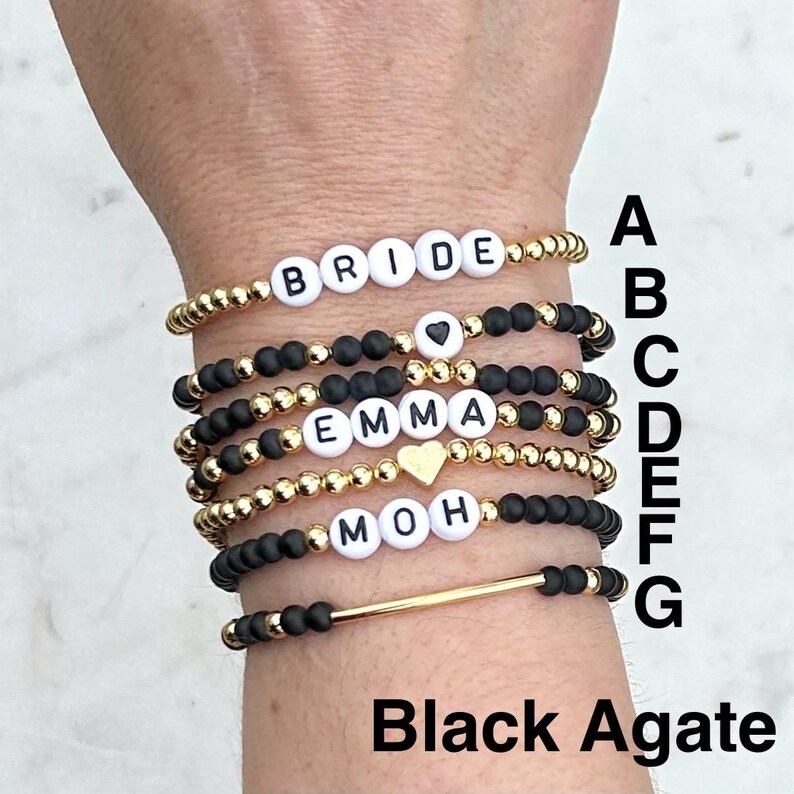 May include: A stack of seven beaded bracelets featuring gold-tone metallic beads, matte black beads, and white letter beads spelling BRIDE, EMMA, and MOH. Some bracelets include heart-shaped beads or a gold-tone bar. The letters A through G are visible.