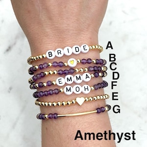 May include: A stack of seven beaded bracelets featuring gold-tone metallic beads and purple amethyst gemstone beads. Some bracelets include white letter beads spelling BRIDE, EMMA, and MOH, while others feature gold heart charms or a gold bar.