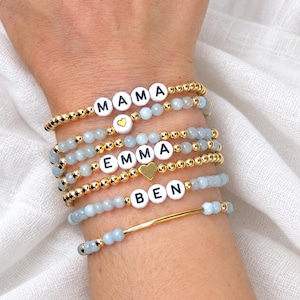 May include: A stack of five beaded bracelets featuring light blue faceted beads and small gold-tone metallic beads. The bracelets include white letter beads spelling MAMA, EMMA, and BEN, along with two small gold-tone heart-shaped charms.
