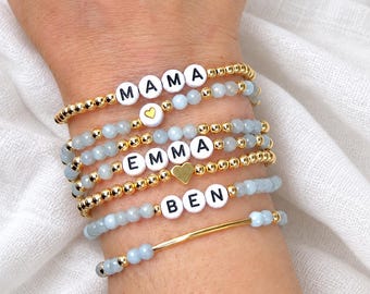 Può includere: Una pila di cinque braccialetti di perline con perline sfaccettate azzurre e piccole perline metalliche dorate. I braccialetti includono perline con lettere bianche che compongono MAMA, EMMA e BEN, e due piccoli ciondoli a cuore dorati.
