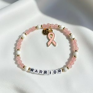 May include: A beaded bracelet featuring light pink and white stone beads, small gold-tone spacers, and white letter beads spelling the word WARRIOR. A gold-tone circular charm and a pink enamel ribbon charm hang from the centre of the bracelet.