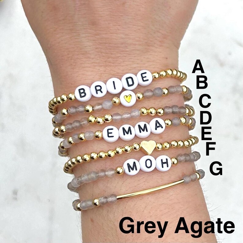 May include: A stack of seven beaded bracelets featuring grey agate beads, gold-tone metallic beads, and white letter beads spelling BRIDE, EMMA, and MOH. Some bracelets include gold-tone heart charms, and one features a smooth gold-tone bar.