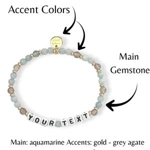 May include: A beaded bracelet featuring aquamarine and grey agate stones, with gold accents. The bracelet includes white letter beads spelling "YOUR TEXT". A small gold charm is attached. The main gemstone is aquamarine.