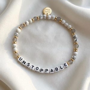 Pode incluir: Uma pulseira de contas com a palavra "UNSTOPPABLE" escrita em contas de letras brancas. A pulseira apresenta contas brancas e cinzentas marmorizadas, contas cinzentas e espaçadores dourados. Um pequeno pingente dourado com as palavras "Simply Stated" pende.