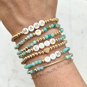 May include: Stack of eight gold bead bracelets with turquoise beads. The bracelets have white beads with gold lettering spelling out the names "MAMA", "ELLA", and "BEN". Two gold heart charms are included.