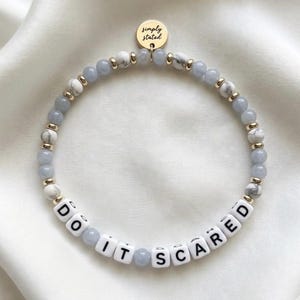 以下が含まれることがあります： 水色と白のマーブル模様のビーズ、ゴールドトーンのスペーサー、DO IT SCAREDと綴られた白い正方形の文字ビーズで構成されたブレスレット。上部にはsimply statedと黒い文字で刻印された丸いゴールドトーンのチャームが付いています。