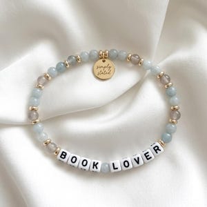Puede incluir: Una pulsera de cuentas con las palabras "BOOK LOVER" deletreadas en cuentas de letras blancas. La pulsera presenta cuentas de color azul claro y gris, detalles dorados y un pequeño dije dorado que dice "simply stated".