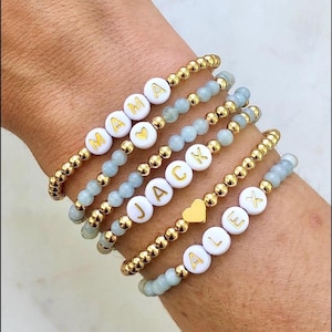 Stacked stretch beaded bracelets with gold-tone and pale-blue beads; white alphabet beads spell names for a personalized kids name bracelet set.