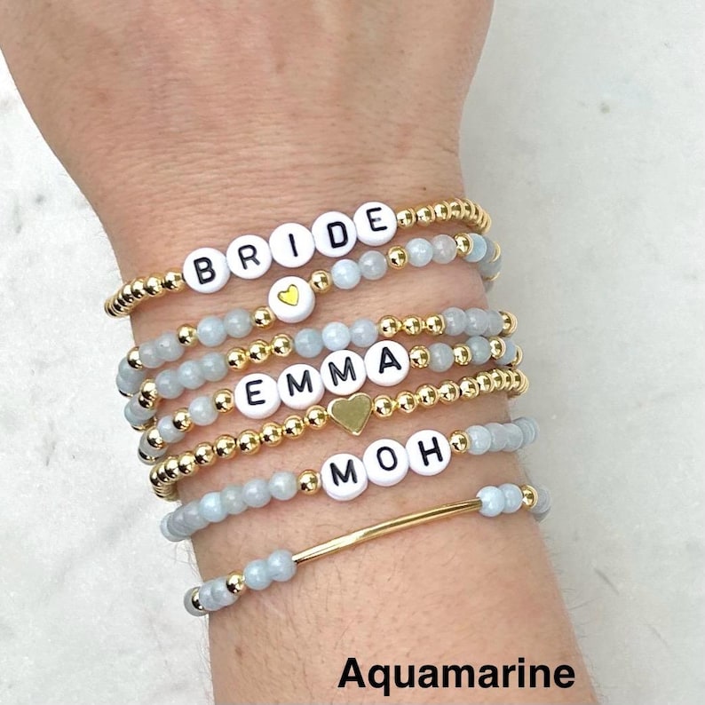 May include: A stack of six stretch bracelets featuring light blue aquamarine beads, small gold-tone metallic beads, and white letter beads spelling BRIDE, EMMA, and MOH. One bracelet includes a gold-tone heart charm and another a gold-tone bar.