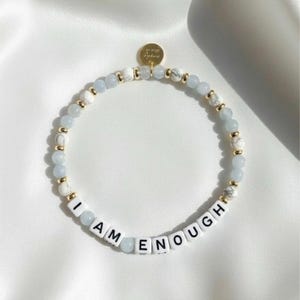 Peut inclure: Un bracelet de perles avec les mots "I AM ENOUGH" épelés en perles de lettres blanches. Le bracelet comporte des perles bleu clair et blanches effet marbre, avec des accents dorés. Une petite breloque dorée est attachée.