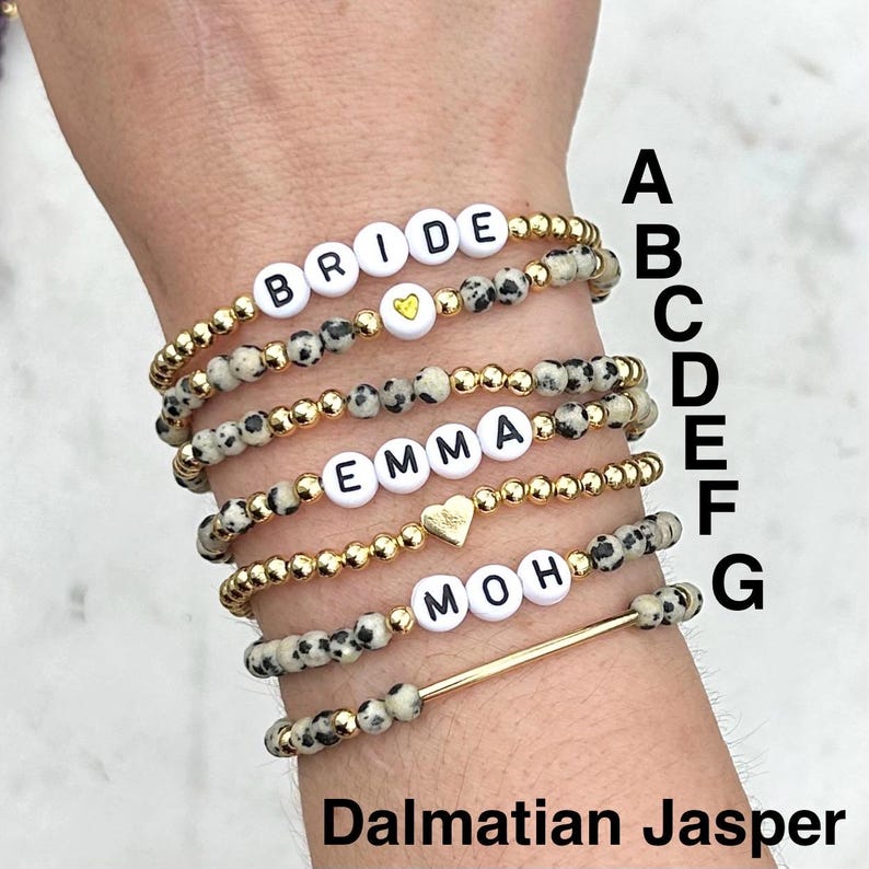 May include: A set of seven beaded bracelets featuring Dalmatian jasper stones, gold-toned metallic beads, and white letter beads spelling BRIDE, EMMA, and MOH. Some bracelets include small gold heart charms or a gold bar. Labeled A through G.