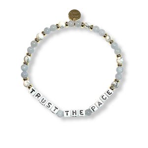 May include: A beaded bracelet featuring a mix of light blue, white, and gold-colored beads. The bracelet includes white square beads spelling out "TRUST THE PACE". A small gold charm is attached. The bracelet is designed for everyday wear.