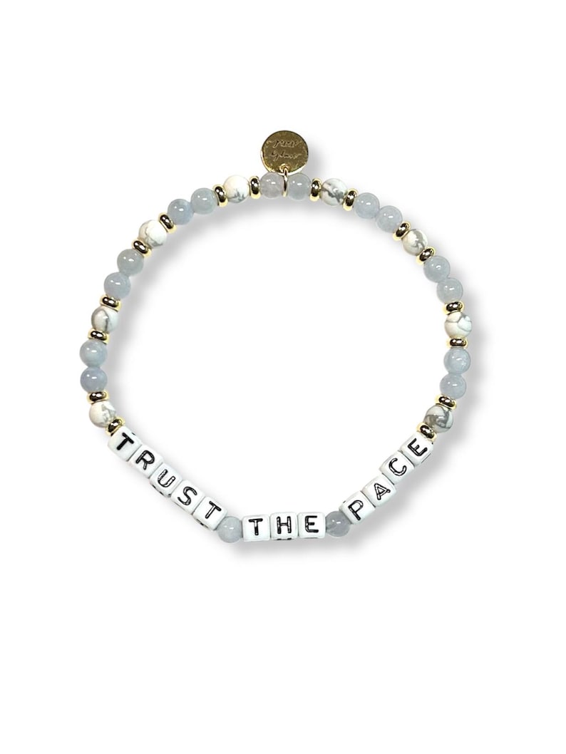 May include: A beaded bracelet featuring a mix of light blue, white, and gold-colored beads. The bracelet includes white square beads spelling out "TRUST THE PACE". A small gold charm is attached. The bracelet is designed for everyday wear.